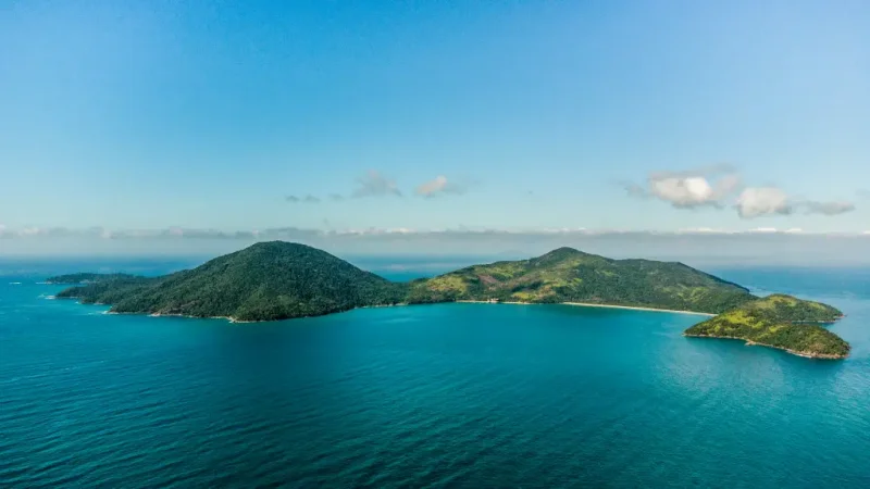 Vista aérea de ilha, coberta por vegetação e cercada por mar de cor azulada.